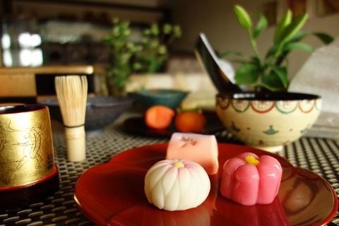 A close-up shows wagashi sweets on a red plate, with a golden cup, tea whisk, fruit, and a bowl with a plant in the background.