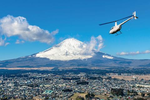 A white helicopter flies past Mount Fuji overlooking a Japanese city.