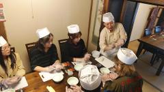 Five people wearing chef's hats and plastic gloves are gathered around a wooden table, learning to make sushi from an instructor.