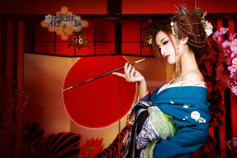 A woman in a teal kimono, styled as a geisha, holds a long pipe against a backdrop featuring a large red circle and Japanese lanterns.