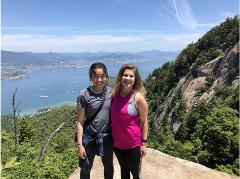 Two women pose for a photo on a mountain, with a city and waterway visible in the background on a bright day.