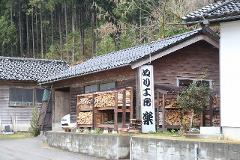 A rustic wooden building, likely a workshop, sits beside a forest, with stacks of wood and a sign in Japanese.