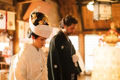 A bride in traditional Japanese wedding attire and a groom in a dark kimono stand together at a wedding ceremony.