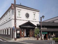 A light-colored, two-story building with a dark-tiled roof and an ornate entrance stands beside a smaller, traditional-looking shop, both viewed from a street with a lamppost in the foreground under a clear blue sky.