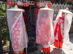 Three women dressed in traditional Japanese clothing, wearing wide-brimmed hats with sheer veils, stand near a temple adorned with various charms and plaques.