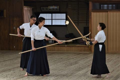 Three people are practicing Kendo, two holding wooden rods facing one woman who holds a wooden sword and padded gloves, all barefoot.