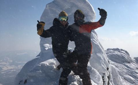 Two people are celebrating on a snow-covered mountain peak, with one giving a thumbs up while standing in front of a snow-covered rock formation.