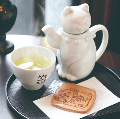 A white ceramic teapot shaped like a lucky cat sits on a tray with a cup of tea and a decorative cookie.