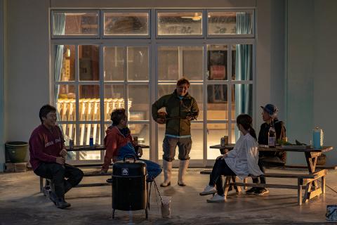 A group of people are gathered outside, sitting on picnic tables and benches, with one man standing and playing a musical instrument, likely enjoying a "Yuntaku Social Exchange and BBQ Plan" on Iriomote Island.