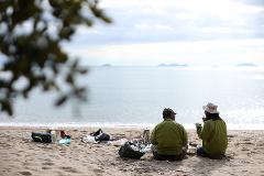 Two people are enjoying a picnic on a sandy beach, sitting and eating with a view of the calm sea and distant islands, with picnic supplies and blurred foliage in the foreground.