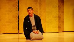 A Japanese man in traditional attire sits cross-legged on a wooden floor, in front of a golden screen, speaking with his hands clasped.