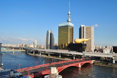 A vibrant cityscape featuring the Tokyo Skytree, Asahi Beer Hall with its golden flame, and a red bridge over a river under a clear blue sky.