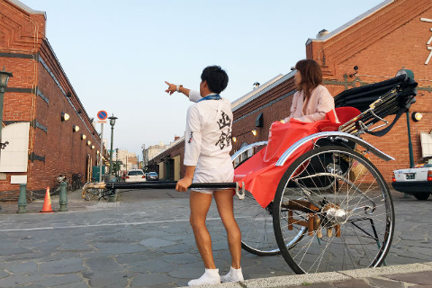A rickshaw puller points the way for a female passenger in front of brick buildings on a cobblestone street.