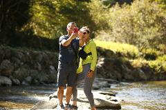 A man and a woman take a selfie with a red phone while standing on rocks in a river with lush green trees in the background.