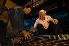A woman in traditional Japanese attire plays a koto while an older man watches attentively, suggesting a cultural exchange.