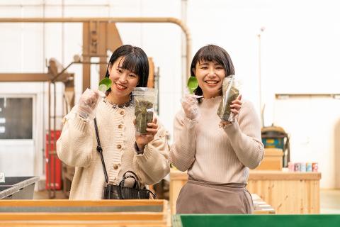 Shizuoka Tea Farm Tour with Blending Experience – 1 Day Japan Trip Two young women in a tea farm hold up bags of tea leaves and scoops, smiling at the camera.