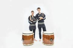 Two performers in traditional Japanese attire stand with arms crossed and drumsticks in hand, beside two large drums in a studio setting.