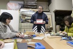 A craftsman shows off a mini tatami while two women work on their own projects at a table.