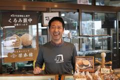 A smiling Japanese man stands behind a counter in a shop, with traditional sweets and signs visible in the background.