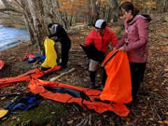 People are preparing gear on a rocky, leaf-strewn shore of a lake next to a forest in autumn.