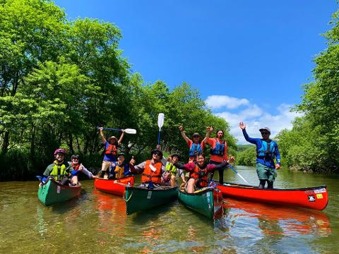 A group of people in canoes are enjoying a sunny day on the Shiodomari River, raising their arms and paddles in celebration.