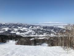A scenic view of a snowy mountain landscape with a ski slope in the foreground and distant mountains under a clear blue sky.