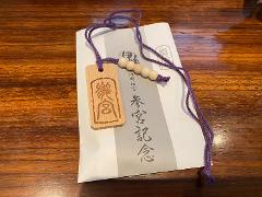 A wooden amulet with purple string and beads rests on a white paper with Japanese text, on a wooden surface.