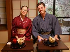 A smiling couple in kimonos sits at tables with tea ceremony sets in a traditional Japanese room with a garden view.