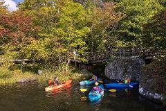 Visit the Spring Water Flowing into Lake Shirakaba and Experience the Origins of Our Connection with Nature – Private Guided Canoe Tour People kayak on a serene lake surrounded by vibrant autumn foliage, with a wooden bridge and rocky boulders framing the picturesque scene.