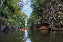 A person kayaks through a narrow river surrounded by lush green cliffs.