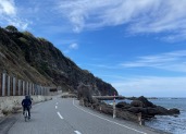 A cyclist rides along a scenic coastal road with a steep, vegetated cliff on one side and the ocean on the other, under a bright blue sky with scattered clouds.