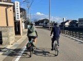 Two people cycle down a paved road alongside a canal under a clear blue sky, with buildings on their left and trees and mountains in the distance.