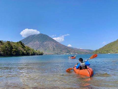 Two people are kayaking on a calm lake with a mountain in the background under a clear blue sky.