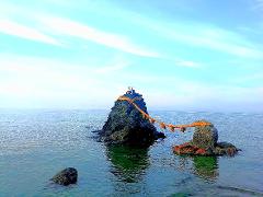 Two rocky islets in the ocean are connected by a decorative rope, with a small torii gate on the larger islet under a bright sky.