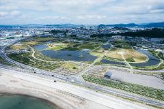 An aerial view shows a coastal park with ponds, roads, and distant mountains, suggesting a site of recovery and remembrance.