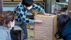 Visit Nikko's Craftsmanship Workshop to Experience Historic Techniques A craftsman demonstrates detailed wood carving techniques on a small chest of drawers to two onlookers in a workshop.