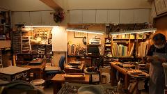 A craftsman, wearing a mask and apron, stands in a workshop filled with tools, materials, and shelves of supplies, examining papers.