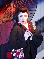 A person in traditional Japanese attire, with white makeup and a black kimono, holds a red and black umbrella while looking intently at the camera.