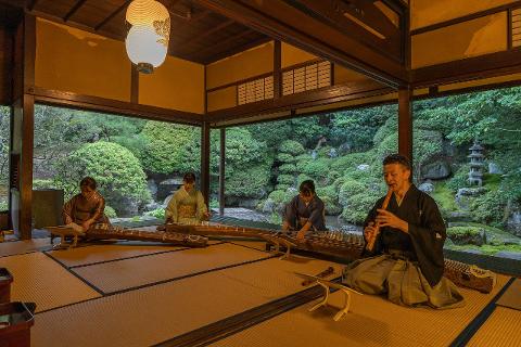 Four musicians in traditional Japanese attire perform koto and shakuhachi music in a room overlooking a serene garden.