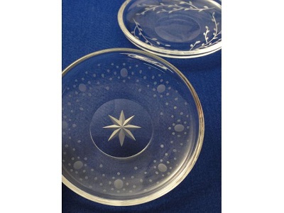 Two clear glass plates with etched designs lay on a blue surface; one features a starburst and dot pattern, the other a stylized floral motif.
