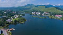 An aerial view shows a serene lake surrounded by lush green hills and scattered buildings, with a road winding through the landscape and several boats on the water.