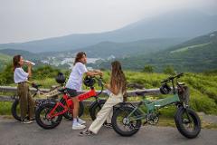 Three young women with their electric bikes enjoy a scenic view of a lake and mountains on a cloudy day.