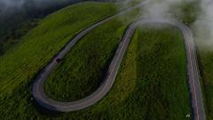 An aerial view shows a winding road on a lush green hillside, shrouded in mist, with a red car driving on it.