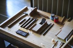 A collection of woodworking tools, including chisels, a plane, a hammer, and a brush, are neatly arranged on a wooden surface, ready for use.