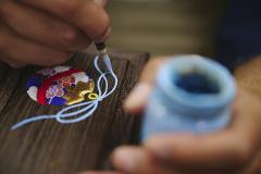 A close-up shot captures a craftsman's hand delicately applying a blue substance with a brush to a handcrafted object, showcasing Japanese artistry.