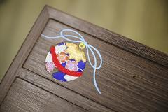 A close-up of a traditional Japanese kiri wood inlaid box features a decorative fabric ornament resembling a round fan with floral patterns, tied with a light blue string and a small yellow loop.