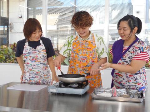 Três mulheres vestindo aventais estão em volta do balcão da cozinha preparando comida em uma panela.