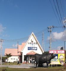 Um restaurante com uma estátua de uma vaca preta na frente e uma placa com texto em japonês que diz &quot;Okinawa Kokuto (açúcar mascavo) Yamami Izumi Restaurant Suppon Kan&quot;, com um céu azul ao fundo.