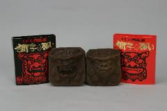A imagem mostra duas estátuas marrons, quadradas, semelhantes a leões, com entalhes detalhados de rostos, sentadas entre uma caixa preta e uma vermelha, ambas adornadas com texto em japonês e um desenho de leão vermelho.