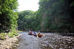 Two canoes with people and a dog paddle down a rocky river flanked by lush, green trees and a cliff face in Jozankei, Hokkaido.